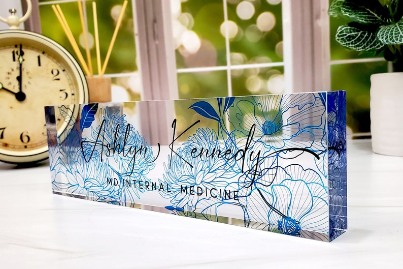 May include: A clear acrylic desk nameplate featuring a floral design in shades of blue. The name "Ashlyn Kennedy" is written in elegant script, with "MD INTERNAL MEDICINE" below. The nameplate is rectangular and stands upright.