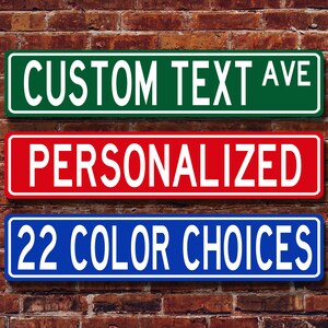 May include: Three rectangular street signs with white text on a brick background. The top sign is green and reads "CUSTOM TEXT AVE". The middle sign is red and says "PERSONALIZED". The bottom sign is blue and says "22 COLOUR CHOICES".