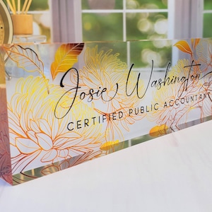 May include: Clear acrylic desk nameplate with floral design in yellow and orange. The nameplate features the name "Josie Washington" and the title "Certified Public Accountant" in black script.