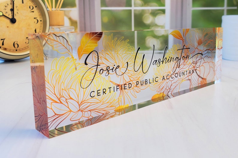 May include: Clear acrylic desk nameplate with floral design in yellow and orange. The nameplate features the name "Josie Washington" and the title "Certified Public Accountant" in black script.