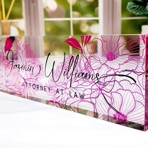 May include: A clear acrylic desk nameplate with a floral design in shades of pink and white. The nameplate features the name "Jasmin Williams" and the title "ATTORNEY AT LAW" in black script.