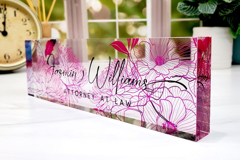 May include: A clear acrylic desk nameplate with a floral design in shades of pink and white. The nameplate features the name "Jasmin Williams" and the title "ATTORNEY AT LAW" in black script.