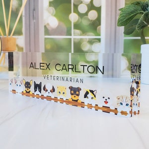 Peut inclure: Une plaque de bureau en acrylique transparente avec le texte "ALEX CARLTON VETERINARIAN" et une rang&eacute;e d'animaux de dessin anim&eacute;, dont des chiens et des chats, qui regardent par-dessus une cl&ocirc;ture en bois.