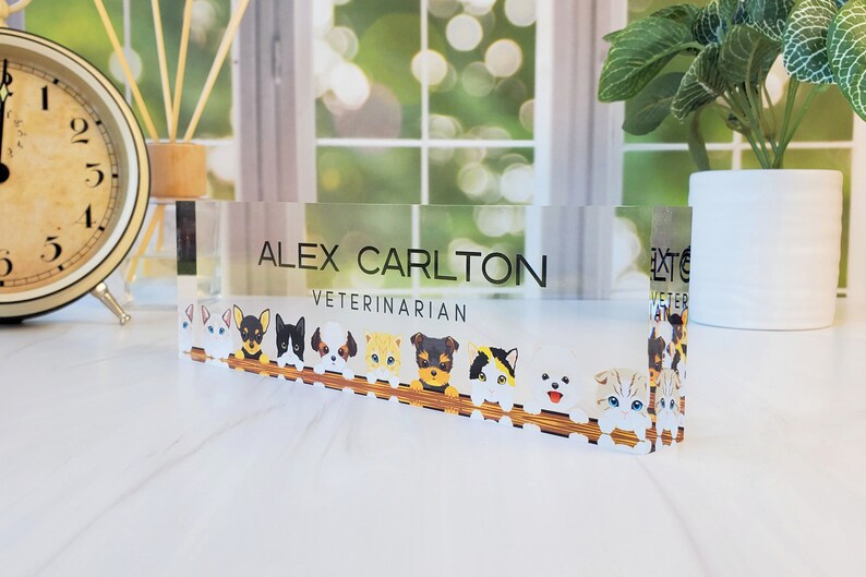 Peut inclure: Une plaque de bureau en acrylique transparente avec le texte "ALEX CARLTON VETERINARIAN" et une rang&eacute;e d'animaux de dessin anim&eacute;, dont des chiens et des chats, qui regardent par-dessus une cl&ocirc;ture en bois.