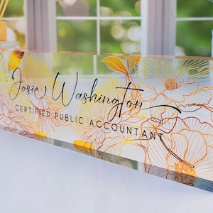 May include: A clear acrylic desk nameplate with gold and orange floral patterns. The name "Josie Washington" and the title "Certified Public Accountant" are written in black script. This rectangular nameplate is a decorative office accessory, suitable for a desk.