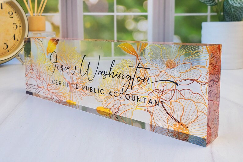 May include: A clear acrylic desk nameplate with gold and orange floral patterns. The name "Josie Washington" and the title "Certified Public Accountant" are written in black script. This rectangular nameplate is a decorative office accessory, suitable for a desk.