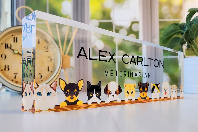 Peut inclure: Une plaque de bureau en acrylique transparente avec le texte "ALEX CARLTON VETERINARIAN" en lettres noires. La plaque pr&eacute;sente une rang&eacute;e d'animaux de dessin anim&eacute;, dont des chiens et des chats, qui regardent par-dessus une surface en bois.
