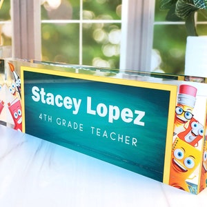 May include: A clear acrylic desk nameplate with a green chalkboard background and yellow border. The nameplate features the text "Stacey Lopez 4th Grade Teacher" and a cartoon image of pencils with faces.