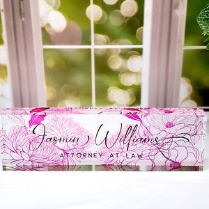 May include: A clear acrylic desk nameplate featuring a pink floral pattern. The name "Jasmin Williams" and the title "ATTORNEY AT LAW" are written in a stylish script.