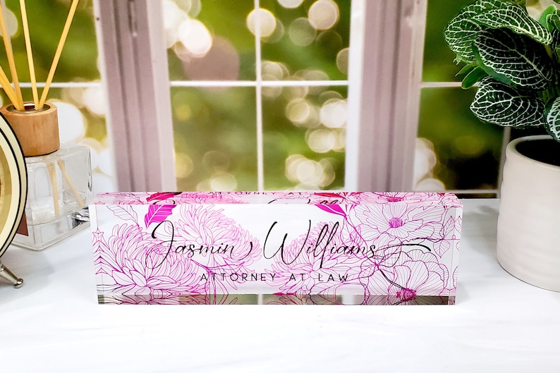 May include: A clear acrylic desk nameplate featuring a pink floral pattern. The name "Jasmin Williams" and the title "ATTORNEY AT LAW" are written in a stylish script.
