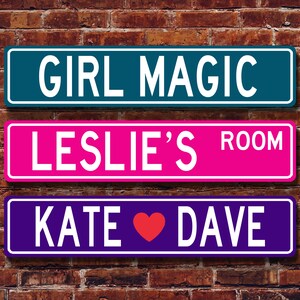 May include: Three rectangular signs with white text against a brick wall. The top sign is teal and reads "GIRL MAGIC." The middle sign is pink and says "LESLIE'S ROOM." The bottom sign is purple and says "KATE ❤ DAVE."