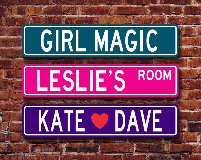 May include: Three rectangular signs with white text against a brick wall. The top sign is teal and reads "GIRL MAGIC." The middle sign is pink and says "LESLIE'S ROOM." The bottom sign is purple and says "KATE ❤ DAVE."