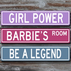 May include: Three rectangular signs with white text on a wooden background. The top sign is lavender and reads "GIRL POWER." The middle sign is pink and reads "BARBIE'S ROOM." The bottom sign is blue and reads "BE A LEGEND."