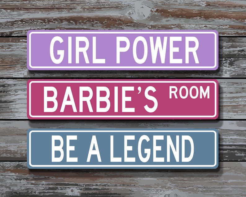 May include: Three rectangular signs with white text on a wooden background. The top sign is lavender and reads "GIRL POWER." The middle sign is pink and reads "BARBIE'S ROOM." The bottom sign is blue and reads "BE A LEGEND."