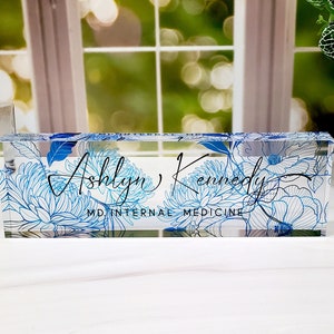 May include: A clear acrylic desk nameplate with the name "Ashlyn Kennedy" in script, and "MD INTERNAL MEDICINE" below. It has blue floral patterns. A decorative office accessory.