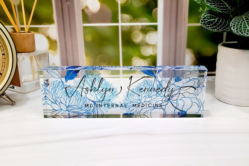 May include: A clear acrylic desk nameplate with the name "Ashlyn Kennedy" in script, and "MD INTERNAL MEDICINE" below. It has blue floral patterns. A decorative office accessory.