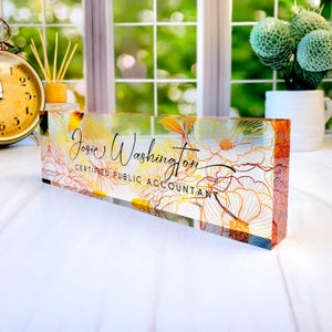 May include: A clear acrylic desk nameplate with floral designs in gold, orange, and white. The nameplate features the name "Josie Washington" and the title "Certified Public Accountant" in black script.