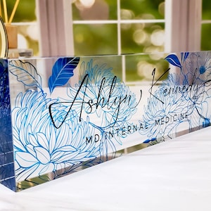 May include: Clear acrylic desk nameplate featuring a blue floral design and the name "Ashlyn Kennedy" in black script. The text "MD INTERNAL MEDICINE" is also visible. The nameplate is rectangular and stands upright.