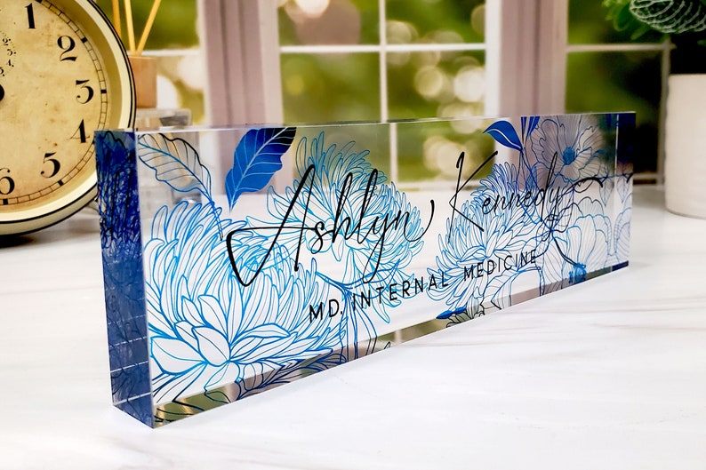 May include: Clear acrylic desk nameplate featuring a blue floral design and the name "Ashlyn Kennedy" in black script. The text "MD INTERNAL MEDICINE" is also visible. The nameplate is rectangular and stands upright.