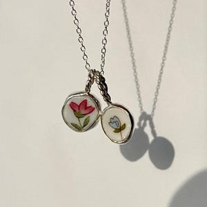 Könnte beinhalten: Eine silberne Kettenkette mit zwei runden Anhängern. Die Anhänger sind weiß mit gemalten Blumendesigns. Ein Anhänger zeigt eine rote Blume und der andere eine blaue Blume.