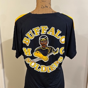 May include: Black t-shirt with yellow shoulder accents. The back features a circular graphic with the words "BUFFALO SOLDIERS" in yellow, surrounding an illustration of a soldier in a yellow uniform holding a sword.