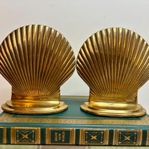 May include: Two gold-colored seashell bookends. Each bookend has a fan-shaped design with vertical lines, sitting on a rectangular base. The bookends are placed on top of a green book with gold lettering and detailing.