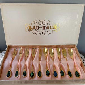 May include: A vintage set of gold-colored flatware, including forks and spoons, presented in a box. The handles feature an abalone inlay. The box lid displays the text "BAU-HAUS" in gold lettering.