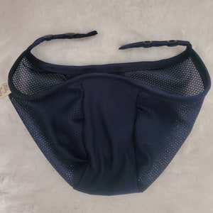 May include: A dark navy blue mesh face covering with a solid center panel and two adjustable straps at the top, each featuring two black plastic snap buttons for secure fastening. The breathable fabric has a fine grid texture throughout the sides.