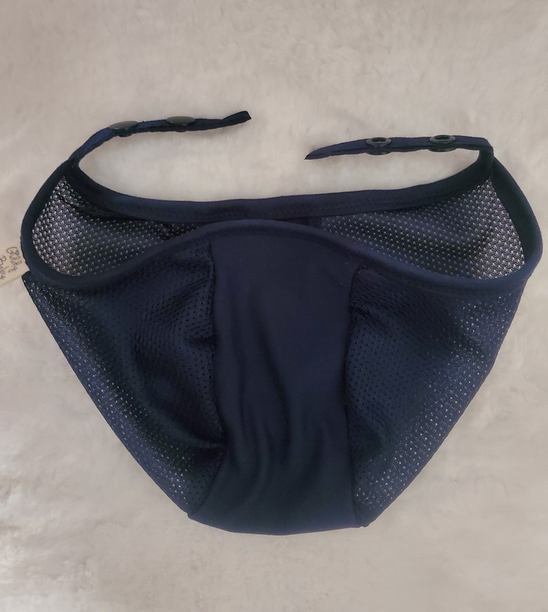 May include: A dark navy blue mesh face covering with a solid center panel and two adjustable straps at the top, each featuring two black plastic snap buttons for secure fastening. The breathable fabric has a fine grid texture throughout the sides.