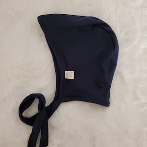 May include: A solid navy blue bonnet with a rounded crown and a curved brim. The headwear features a small white rectangular fabric tag on the side with the text Goldy Baby printed in black. Two long navy blue fabric ties extend from the base.