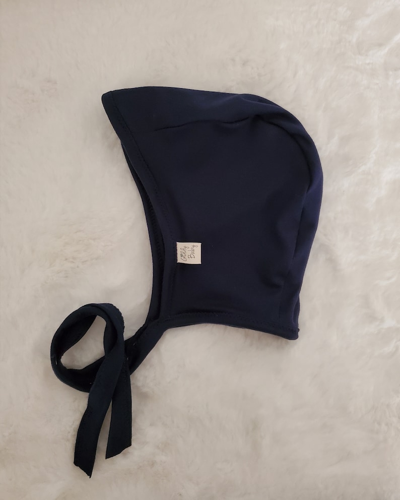 May include: A solid navy blue bonnet with a rounded crown and a curved brim. The headwear features a small white rectangular fabric tag on the side with the text Goldy Baby printed in black. Two long navy blue fabric ties extend from the base.