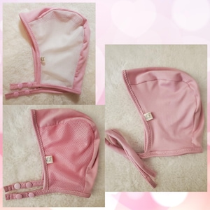 Puede incluir: Gorro de bebé rosa con un panel frontal de malla y cierres de botón a presión. El gorro tiene una tela suave y ligera y una pequeña etiqueta en el lateral. Diseñado para proteger la cabeza del bebé.