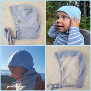 May include: A light blue baby bonnet featuring a smooth exterior, a white mesh interior lining, and two snap closures on the chin strap. A small rectangular fabric tag is sewn onto the side of the bonnet, which is designed to cover the head and ears.