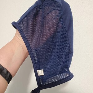 May include: A navy blue bonnet featuring a solid fabric top panel and a semi-sheer mesh side panel. The headwear includes a small white rectangular tag with the text "Goldy Baby" stitched near the bottom edge and two adjustable chin straps.