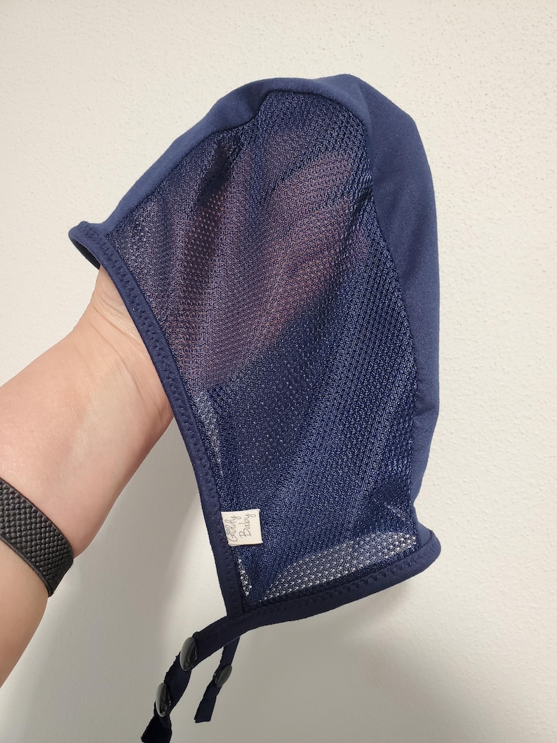 May include: A navy blue bonnet featuring a solid fabric top panel and a semi-sheer mesh side panel. The headwear includes a small white rectangular tag with the text "Goldy Baby" stitched near the bottom edge and two adjustable chin straps.