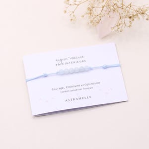 Peut inclure: Un bracelet bleu clair avec cinq perles bleu pâle sur un cordon assorti. Il est présenté sur une carte blanche avec du texte français, dont "aigue-marine" et "ASTRANELLE". Une branche de fleurs séchées est également visible.