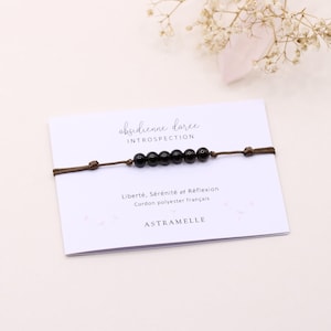 Peut inclure: Un bracelet composé de six petites perles sphériques noires lisses enfilées sur un fin cordon en polyester marron. Le bracelet est présenté sur une carte blanche avec le texte "obsidienne dorée INTROSPECTION" et "Liberté, Sérénité et Réflexion".