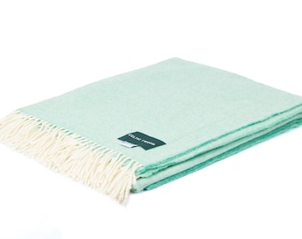 Può includere: Una coperta color verde menta piegata con una trama tessuta e frange color crema lungo il bordo. Un'etichetta rettangolare scura è cucita sulla superficie, riportante il testo bianco CELTIC TWEED in un carattere chiaro senza grazie.