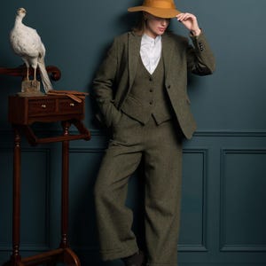 May include: A three-piece olive green tweed suit with a matching hat. The suit includes a blazer, vest, and wide-leg trousers. The person is wearing a white collared shirt and black boots. A decorative wooden stand with a bird is on the left.