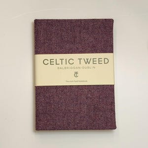 May include: A rectangular notebook covered in a textured purple tweed fabric. A cream-colored paper band wraps around the center, featuring the text CELTIC TWEED, BALBRIGGAN-DUBLIN, a circular logo with the letters CT, and The Irish Field Notebook.
