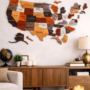 Peut inclure: Une carte en bois des États-Unis, chaque État étant découpé et teinté dans diverses nuances de marron, d'orange et de gris. La carte est fixée sur un mur blanc au-dessus d'un meuble en bois. Un globe, une plante et des bougies sont sur le meuble.