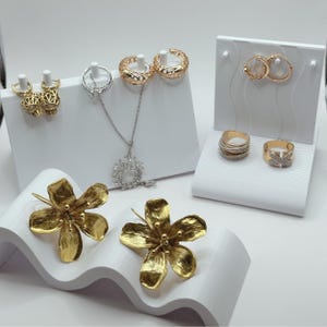 Peut inclure: Un présentoir de bijoux en or et en argent, comprenant des boucles d'oreilles, des bagues et un collier. Les bijoux sont exposés sur des supports blancs à motif ondulé.