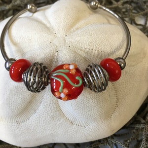May include: A silver-toned bangle bracelet with red glass beads and wire cage accents. The central bead is red with green and white floral designs. The bracelet is displayed on a white, textured surface.