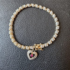 Może przedstawiać: Złota bransoletka z serią małych, przezroczystych kamieni. Z bransoletki zwisa zawieszka w kształcie serca z czerwoną kamieniem. Bransoletka leży na ciemnej, teksturowanej powierzchni.