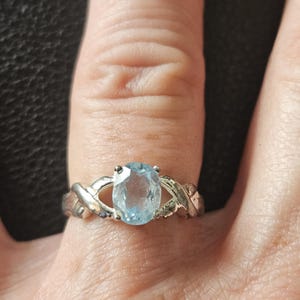 Op de afbeelding: Een zilverkleurige ring met een centrale ovaal geslepen lichtblauwe edelsteen die door vier pootjes wordt vastgehouden. De band heeft een gedraaid, gekruist ontwerp aan beide zijden van de steen, wat een open, verweven uiterlijk creëert.