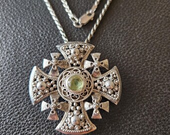 May include: A silver necklace with a large, ornate cross pendant featuring a green gemstone in the center. The cross is decorated with intricate filigree and beading.