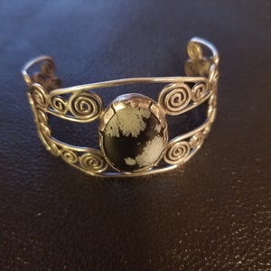 Puede incluir: Una pulsera de plata con una piedra negra y blanca engastada en el centro. La pulsera está hecha de alambre que se retuerce en un patrón de espiral.