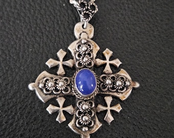Puede incluir: Un collar de plata con un colgante en forma de cruz adornado con una piedra azul en el centro. La cruz está decorada con pequeñas cuentas de plata y cuatro cruces más pequeñas.