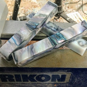 May include: Three rectangular pieces of iridescent, white, blue, and green marbled material. The material is likely a type of resin or plastic. The pieces are stacked on top of each other on a gray surface with the word "RIKON" printed on it.
