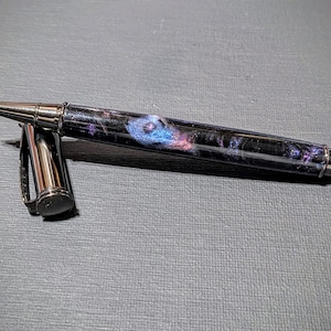 May include: A black and silver pen with a galaxy-themed design. The pen has a black barrel with a silver clip and a silver cap. The galaxy design features swirling colors of blue, pink, and purple.